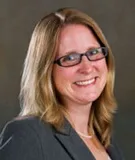 A professional headshot of a woman with long blonde hair, wearing glasses and a gray blazer, smiling. The background is a soft gradient of muted earth tones.