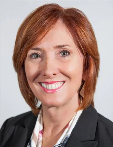 A smiling woman with short auburn hair wearing a black blazer over a light patterned blouse. She is posed against a plain white background.