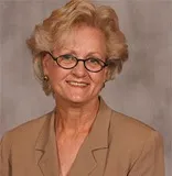 A professional headshot of a woman with blonde hair styled neatly and wearing glasses. She is dressed in a beige suit jacket and is smiling warmly. 