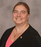 A smiling woman with light brown hair tied back wearing a black blazer over a pink top, accessorized with a gold necklace and a small lapel pin. She is posed against a plain gray background.