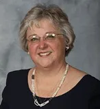 A professional portrait of a woman with short, gray hair, wearing glasses and a navy blue top adorned with a beaded necklace.