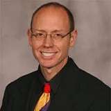 A professional headshot of a man wearing glasses, a black suit, and a multicolored tie with red, yellow, and purple elements.