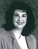 A black-and-white professional portrait of a woman with medium-length, dark hair and bangs. She is wearing a checkered blazer over a white top and smiling softly.