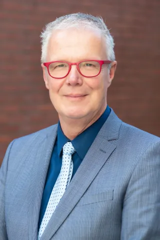 A portrait of a man with short, white hair and fair skin. He is wearing red-framed glasses, a light blue suit jacket, a teal dress shirt, and a patterned tie. 