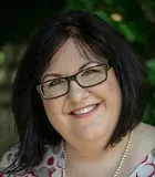 A headshot of a woman with shoulder-length dark hair, wearing glasses and a pearl necklace. She is smiling warmly and is photographed outdoors with greenery in the background.