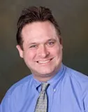 A smiling man with short brown hair wearing a light blue dress shirt and a patterned tie. He is posed against a softly blurred brown and green background.