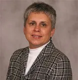 A professional portrait of a woman with short gray hair wearing a checkered blazer over a white turtleneck. She has a confident and composed expression, with a neutral background adding to the formal and polished tone of the image.