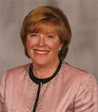 A professional portrait of a woman with short auburn hair, smiling warmly. She is wearing a light pink blazer with a delicate patterned design and a black trim along the collar. 