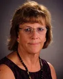 A headshot of a woman with short brown hair wearing glasses, a dark sleeveless top, and a black beaded necklace. She is photographed against a dark background with a neutral expression.