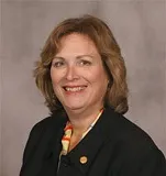 A professional portrait of a woman with shoulder-length light brown hair, wearing a black blazer over a colorful blouse. She is smiling warmly, and the background is a neutral tone, providing a clean and formal look.