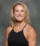 A professional portrait of a woman with blonde hair, wearing a sleeveless black top and a necklace. She is smiling confidently, with a neutral gray background that provides a formal and balanced setting for the photo.