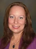 A smiling woman with long reddish-brown hair wearing a purple top and a necklace with a dark pendant. She is posed against a softly blurred dark background.