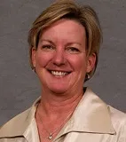 A smiling woman with short blonde hair wearing a beige satin blouse and a necklace with a small pendant. She is posed against a plain gray background.