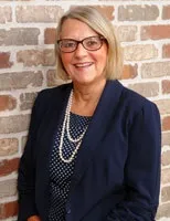 A smiling woman with short blonde hair and glasses wearing a navy blazer over a polka-dotted blouse, accessorized with a pearl necklace. She is posed against a brick wall background.