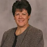 A smiling woman with short dark hair wearing a gray textured blazer over a black top. She is posed against a plain gray background.