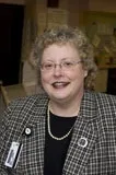 A portrait of a woman with short, curly blonde hair and light skin. She is smiling and wearing glasses, a black and white checkered blazer, a black top, and a pearl necklace. A name badge is visible on her blazer. 