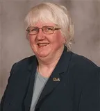 A smiling woman with short white hair and glasses wearing a dark blazer over a light blue top, accessorized with a small lapel pin. She is posed against a plain gray background.