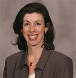 A professional headshot of a woman with shoulder-length black hair. She is smiling brightly, wearing a gray blazer over a white top, accessorized with a red pendant necklace.