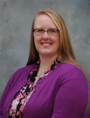 A professional portrait of a smiling woman with long blonde hair. She is wearing glasses, a purple cardigan, and a floral-patterned blouse.