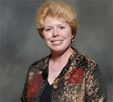 A woman with short, strawberry blonde hair smiles in a professional portrait. She is wearing a patterned black and gold jacket over a dark top.