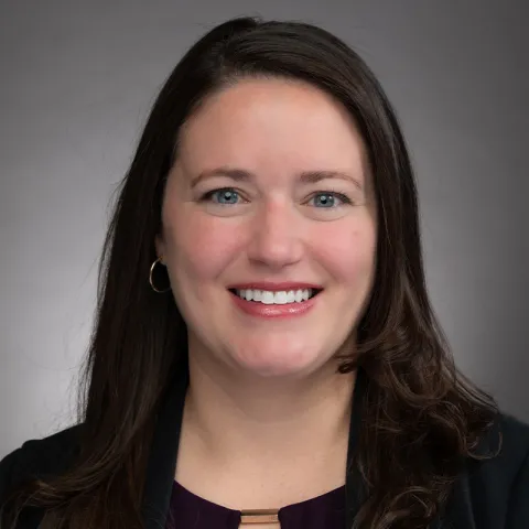 A portrait of a woman with long, straight dark brown hair and light skin. She is smiling brightly and wearing a dark purple blouse with a gold accent at the neckline, paired with a black blazer. She has small hoop earrings. The background is a neutral gray gradient, giving the image a professional and polished appearance. Her expression is warm and approachable.