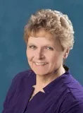 A portrait of a woman with short, curly blonde hair and light skin. She is smiling warmly and wearing a purple blouse.