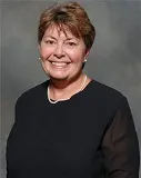 A smiling woman with short brown hair wearing a black top with sheer sleeves, accessorized with a pearl necklace. She is posed against a textured gray background.