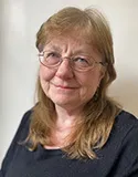 Headshot of a woman with long light brown hair, wearing glasses and a black top, smiling gently.