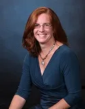 Portrait of a woman with shoulder-length red hair, wearing glasses, a teal dress, and a silver necklace, smiling brightly.