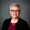 A professional headshot of a woman with short silver-gray hair, wearing glasses, a black cardigan, and a burgundy top.