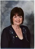 A woman with short dark hair wearing a dark top adorned with decorative embroidery. She is smiling and wearing a gold pin on her left side. The background has a soft, cloudy texture, giving the image a formal portrait look.