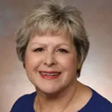 A professional portrait of a woman with short, light grayish-blonde hair, smiling warmly. She is dressed in a vibrant royal blue top and has a subtle makeup look.