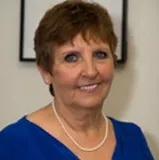 A professional headshot of a smiling woman with short brown hair. She is wearing a blue top and a pearl necklace.