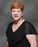 A woman with short red hair wearing a black dress with sheer sleeves and drop earrings. She is posed against a textured gray background.