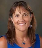 A headshot of a smiling woman with shoulder-length brown hair, wearing a blue sleeveless top and a silver necklace with a blue pendant. She is photographed against a blurred, neutral background.