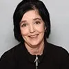 A professional headshot of a woman with short, dark hair styled simply. She is wearing a black top and smiling gently, with a light gray background.