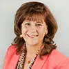 A woman with shoulder-length brown hair smiles in a professional portrait. She is wearing a coral blazer and a patterned blouse, accessorized with earrings and a gold necklace.