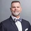 A professional portrait of a man with short gray hair and a neatly trimmed beard. He is wearing a navy blue suit, a white dress shirt, and a patterned bow tie.