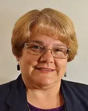 Headshot of a woman with short blonde hair, wearing glasses, a navy blazer, and a purple top, smiling softly.