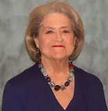 An older woman with styled gray hair and red lipstick wearing a dark blue blouse and a multicolored beaded necklace. She is posed against a textured gray background.