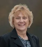 A smiling woman with short blonde hair wearing a black blazer over a patterned blouse. She is posed against an outdoor, softly blurred background.