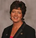 A professional portrait of a woman with short, dark brown hair styled neatly. She is smiling warmly and wearing a black blazer over a pink blouse, accessorized with a delicate necklace.