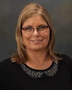 Headshot of a woman with shoulder-length blonde hair, wearing glasses, a black top with an embellished collar, and a necklace, smiling softly.