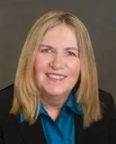 A smiling woman with straight blonde hair wearing a black blazer over a blue collared shirt. She is posed against a plain gray background.