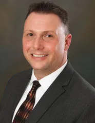 A smiling man with short dark hair wearing a gray pinstripe suit, white shirt, and patterned tie. He is posed against a softly blurred dark background.