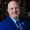 A professional headshot of a man with a bald head, dressed in a sharp blue suit, light blue shirt, and a striped tie.