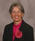 A woman with short, gray hair smiles in a professional portrait. She is wearing glasses, a black pinstripe blazer, and a bright pink blouse accessorized with a large pink flower.