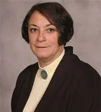 A woman with short, dark brown hair poses for a professional portrait. She is wearing a dark blazer over a light cream blouse and has a decorative brooch at her neckline.