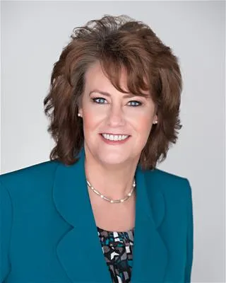 A professional portrait of a woman with voluminous, medium-length brown hair. She is wearing a teal blazer over a patterned blouse and a pearl necklace.