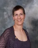 A portrait of a woman with short, dark hair and light skin. She is smiling and wearing glasses, a purple crocheted cardigan, and a matching top. A small pin is visible on her cardigan.